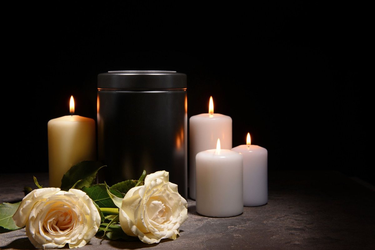 Urna cineraria nera tra candele accese e rose bianche su fondo scuro