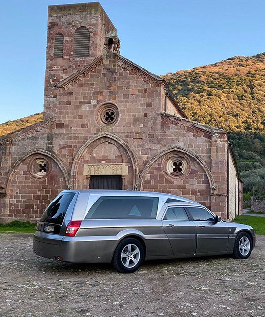 Carro funebre dell'Agenzia Funebre Pinna fotografato da dietro sul sagrato in terra di una chiesa in trachite a Bosa